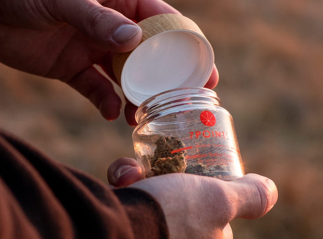 Hands open a small 7Points branded glass jar with a wood lid containing cannabis flower.