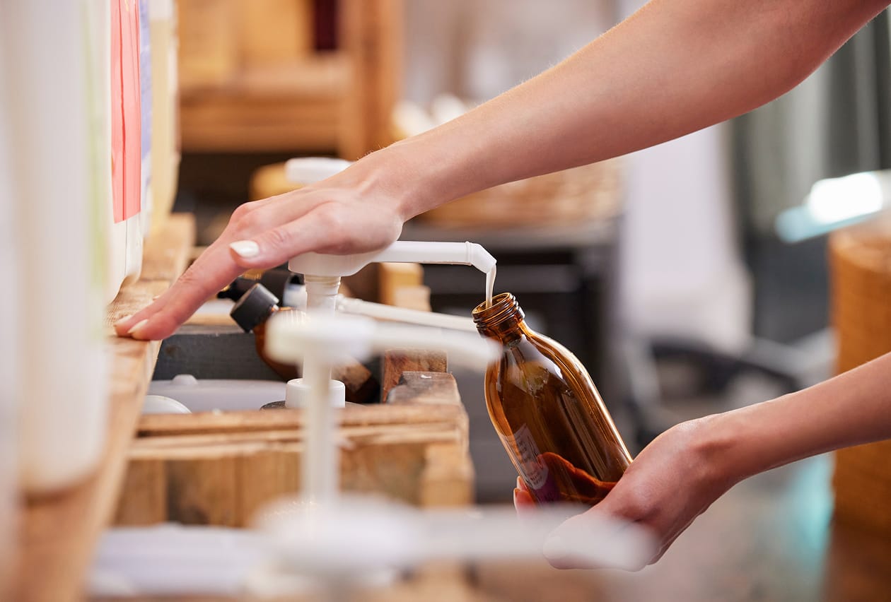 A person refills an amber glass bottle at a bulk liquid dispenser station in a zero-waste store.