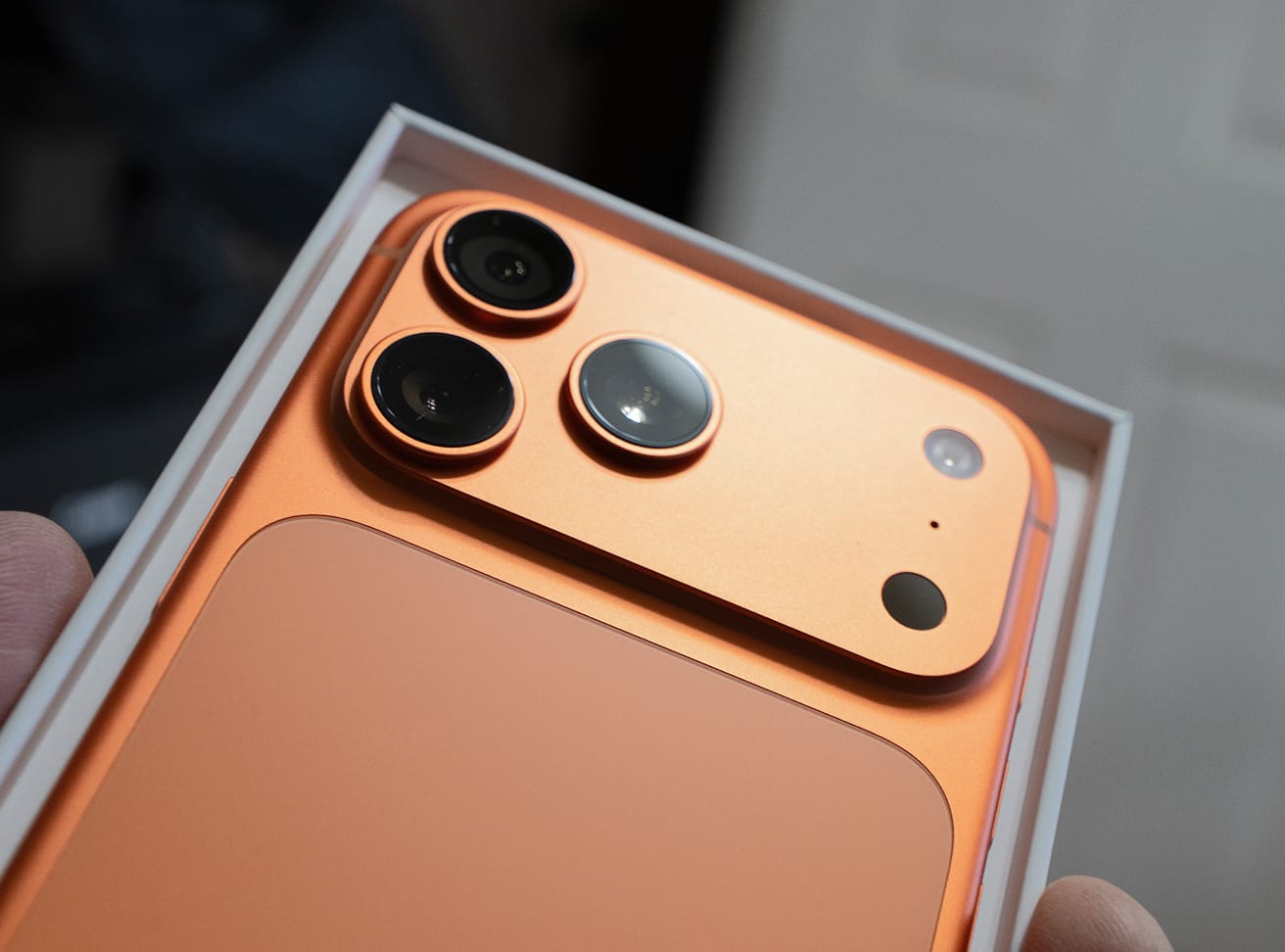 Close-up of an orange smartphone with triple camera system being unboxed from white retail packaging.
