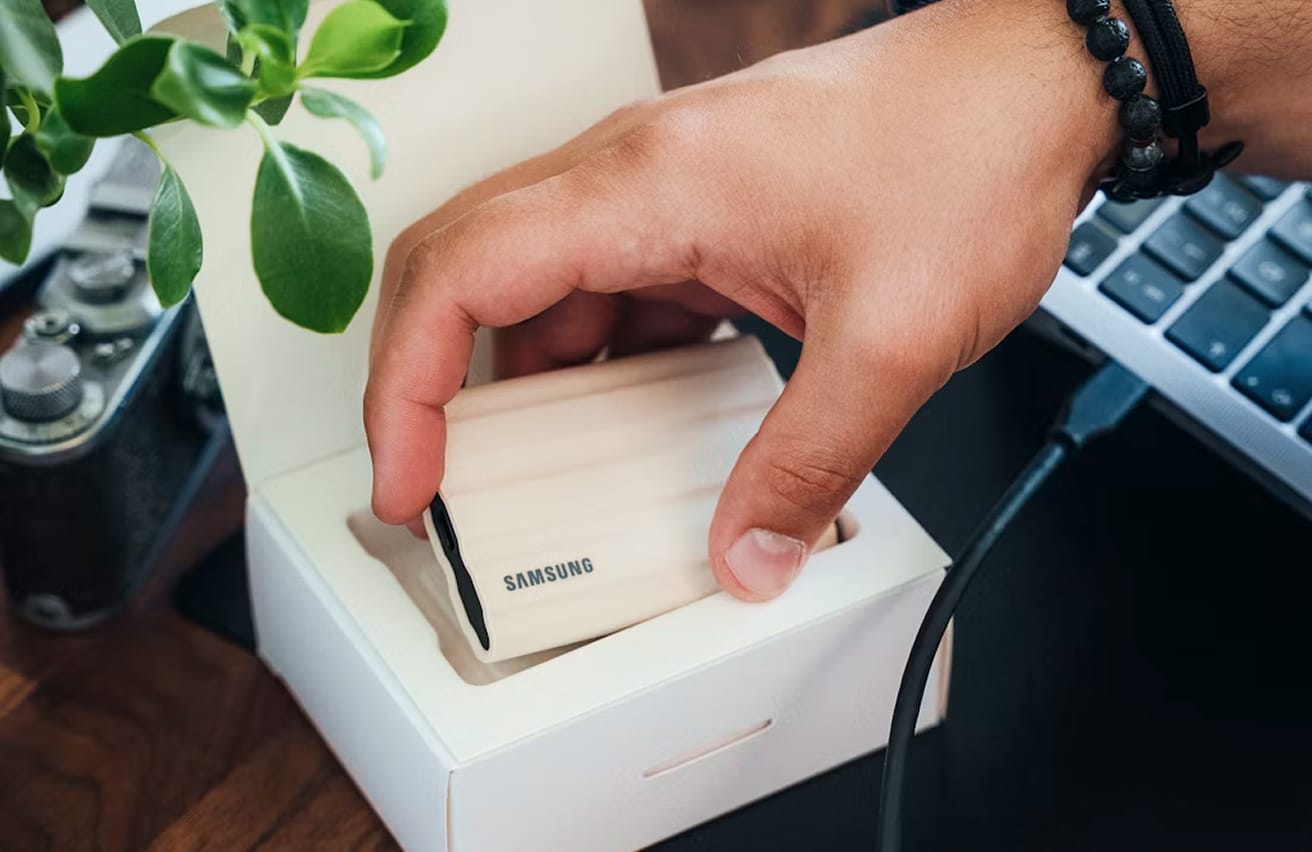 Person removing a beige Samsung portable SSD from its white retail box on a desk.