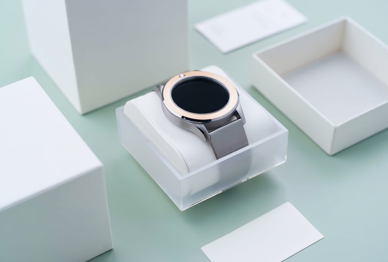 Silver and rose gold smartwatch displayed on a cushioned stand surrounded by minimal white packaging components.