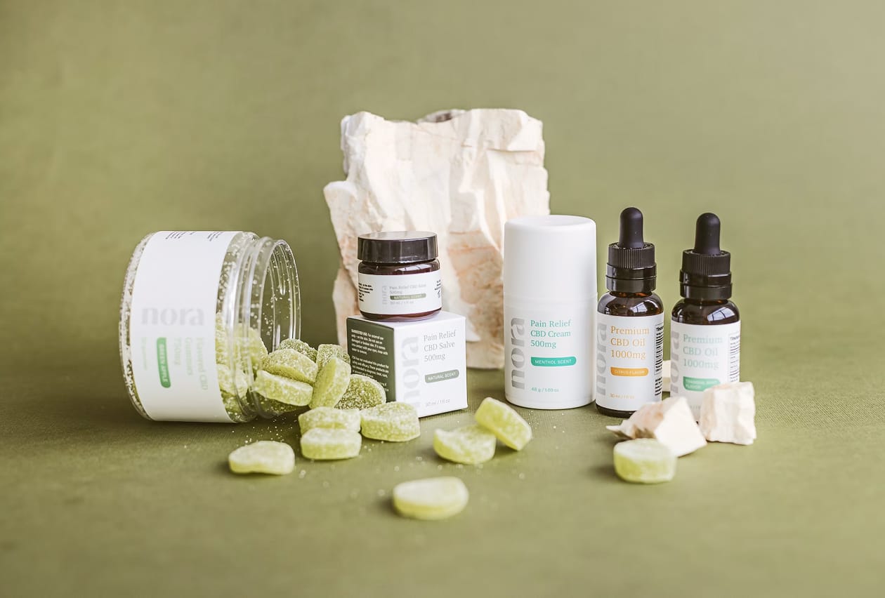 Assortment of Nora brand CBD products including gummies, salve, cream, and tincture bottles on green background.