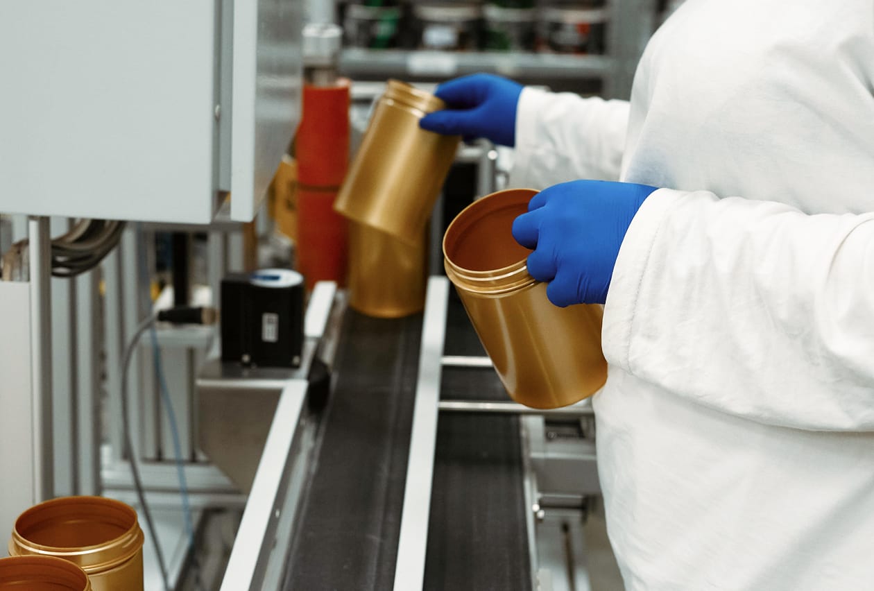 Gloved worker in white lab coat inspecting gold supplement containers on a manufacturing conveyor belt.